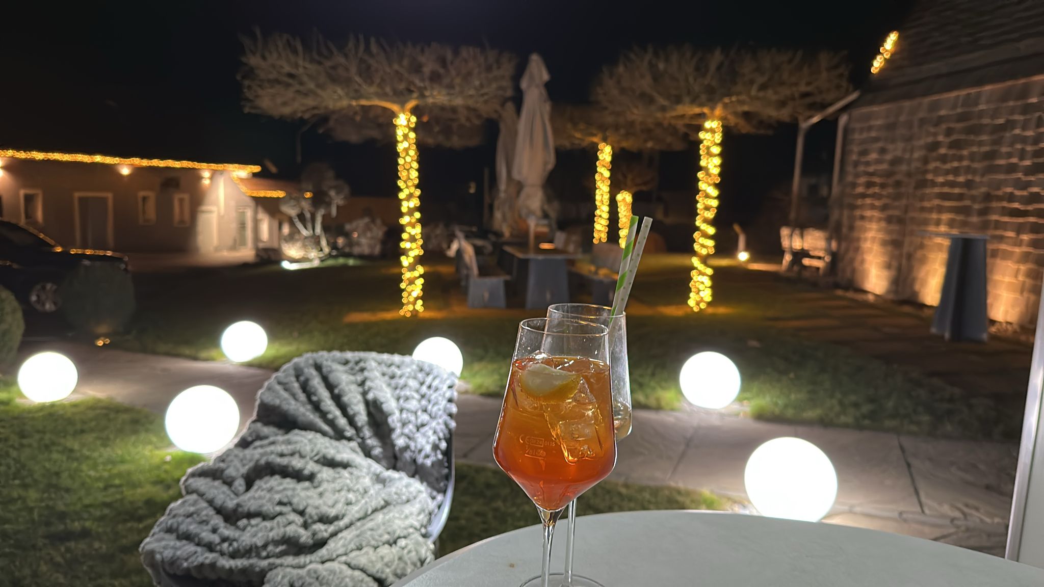 Aperitif-Gläser im Vordergrund, dahinter abendliche Gartenstimmung mit Lichterketten und Kugelleuchten.