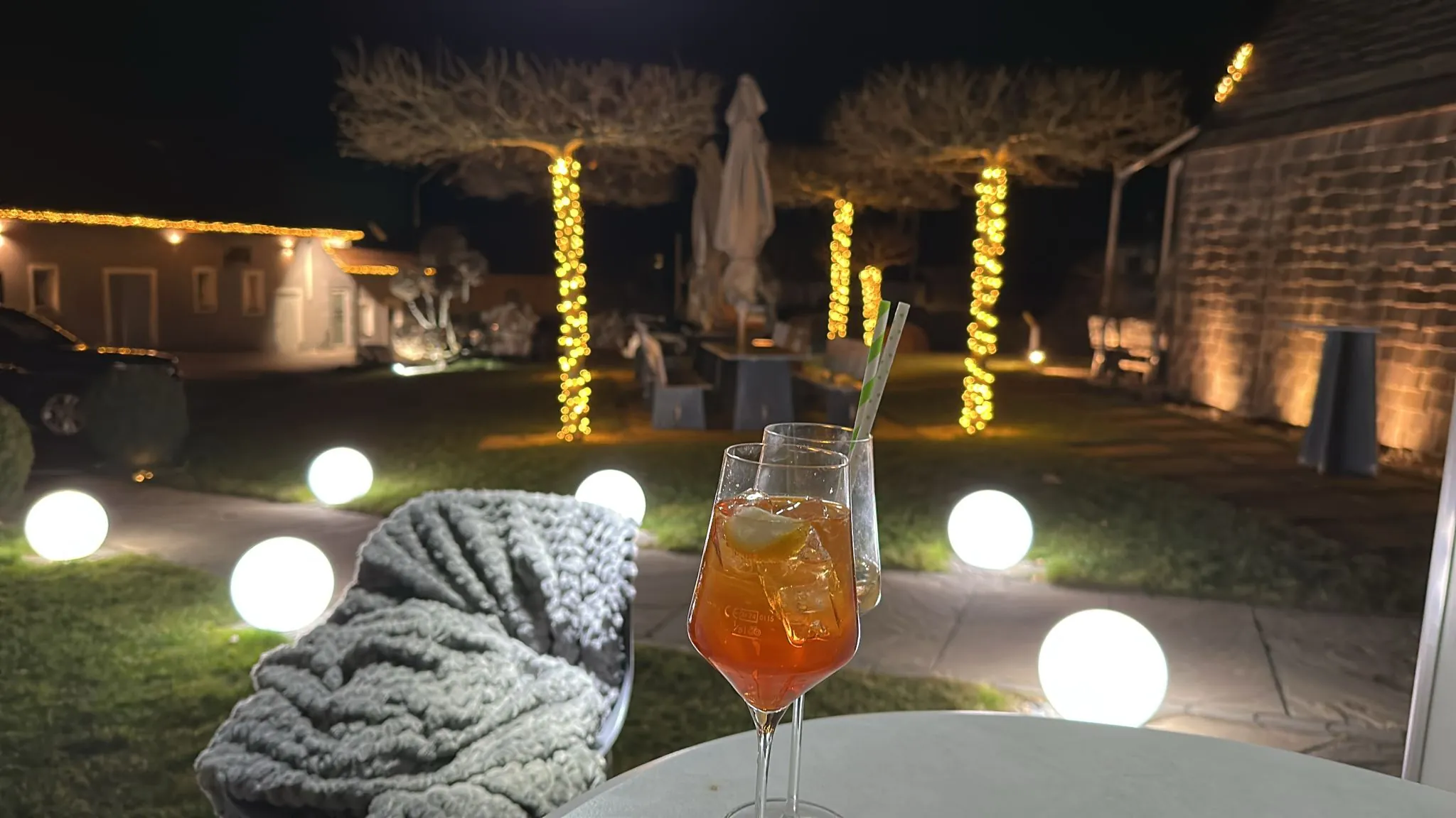 Aperitif-Gläser im Vordergrund, dahinter abendliche Gartenstimmung mit Lichterketten und Kugelleuchten.